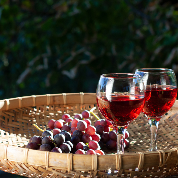 Two glasses of red wine alongside a bunch of grapes in a woven basket, set against a blurred natural background.